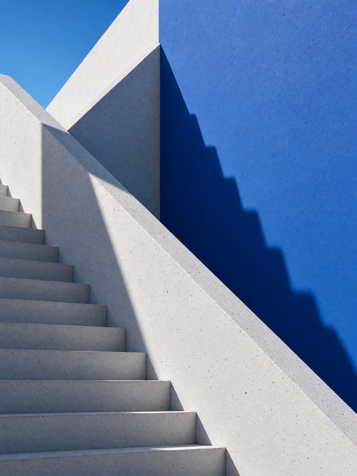A white staircase beside a blue wall, forming diagonal patterns of sunlight and shadow.